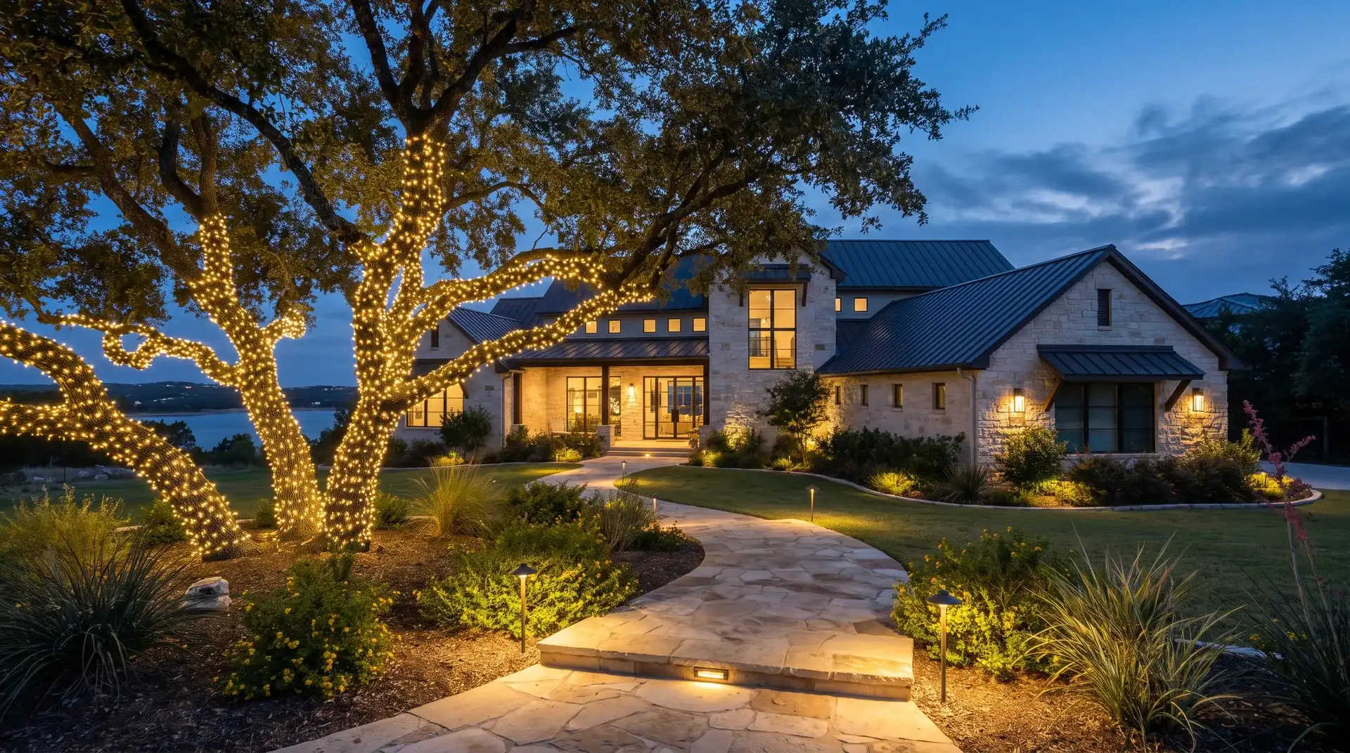 Live oak wrapped in Christmas lights with lit landscaping at a Lake Travis hillside estate in Lakeway, TX