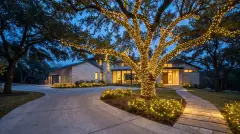 Live oak wrapped in Christmas lights with lit landscaping at a wooded hillside home in West Lake Hills, TX